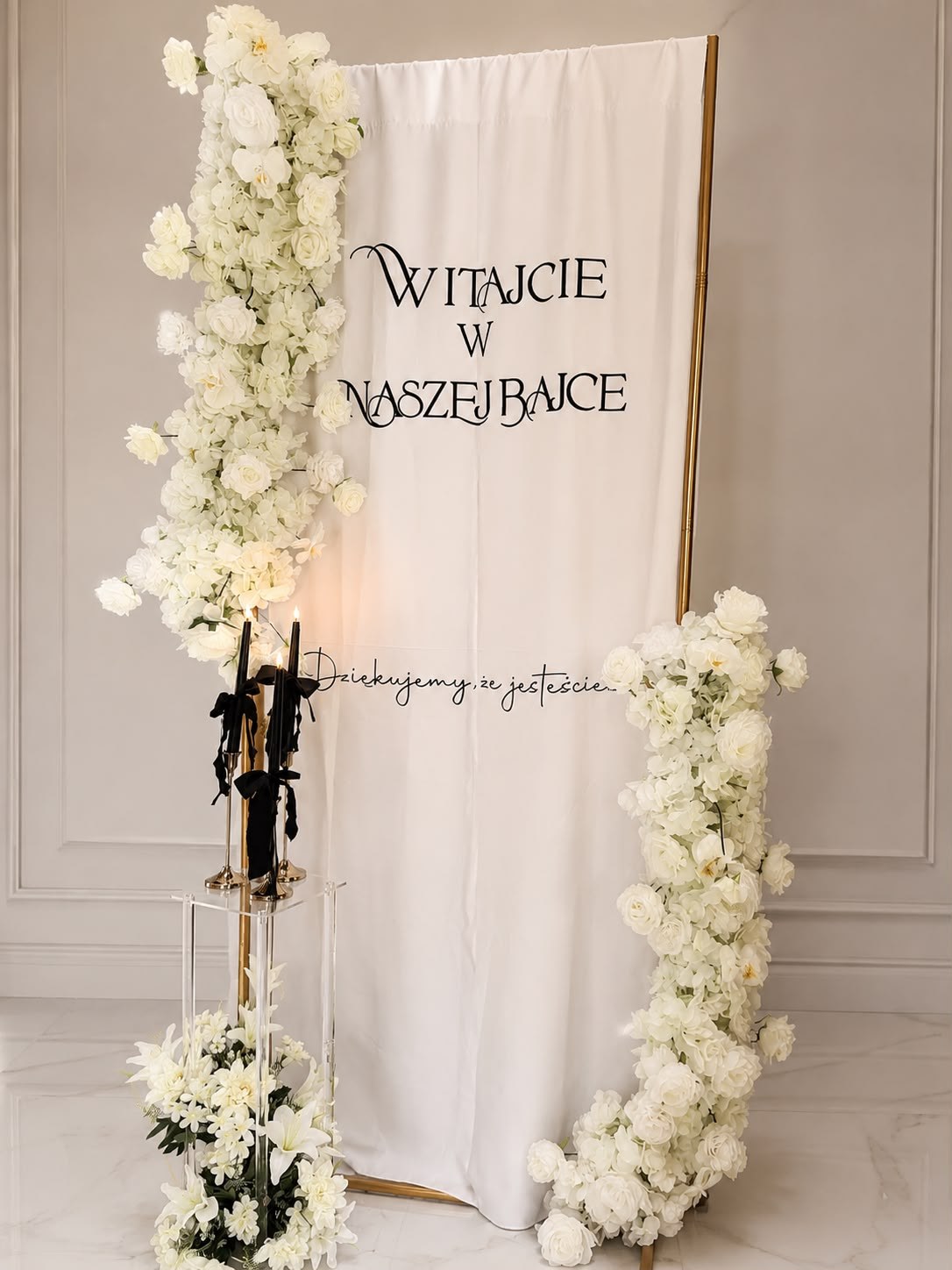 Biała ścianka powitalna „Witajcie w naszej bajce” z białymi kwiatami i świecami